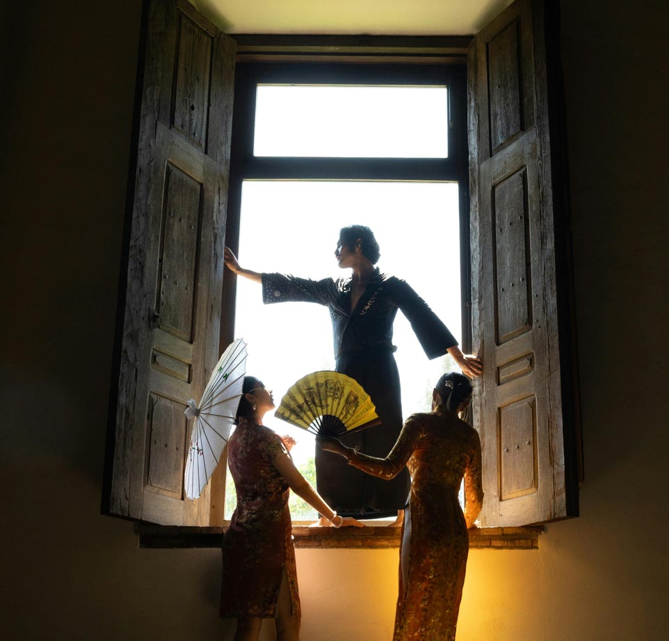Three women in elegant attire with fans and parasol, framed in dramatic window light, creating an artistic silhouette.